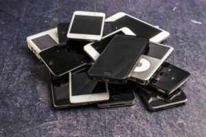 **Alt Text:** A pile of old iPhones and iPods on a dark textured surface.