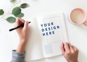 Alt text: A person writes in a notebook that has the words "Your Design Here" printed on the page, with a nearby mug and green leaves on the table.