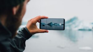 **Alt text:** A person holds a smartphone up to capture a photo of icebergs on a calm, foggy body of water.