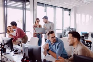 Alt text: A group of people collaborating in a modern office with two discussing over a laptop, and others talking at desks with computers.
