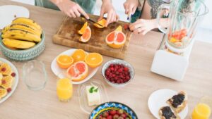 Alt text: Two people cutting fruits on a wooden board surrounded by bananas, oranges, raspberries, juice, and a blender on a kitchen countertop.