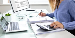 **Alt Text:** A woman in a blue shirt uses a calculator and writes on documents at a desk with a laptop and computer monitor displaying a spreadsheet.