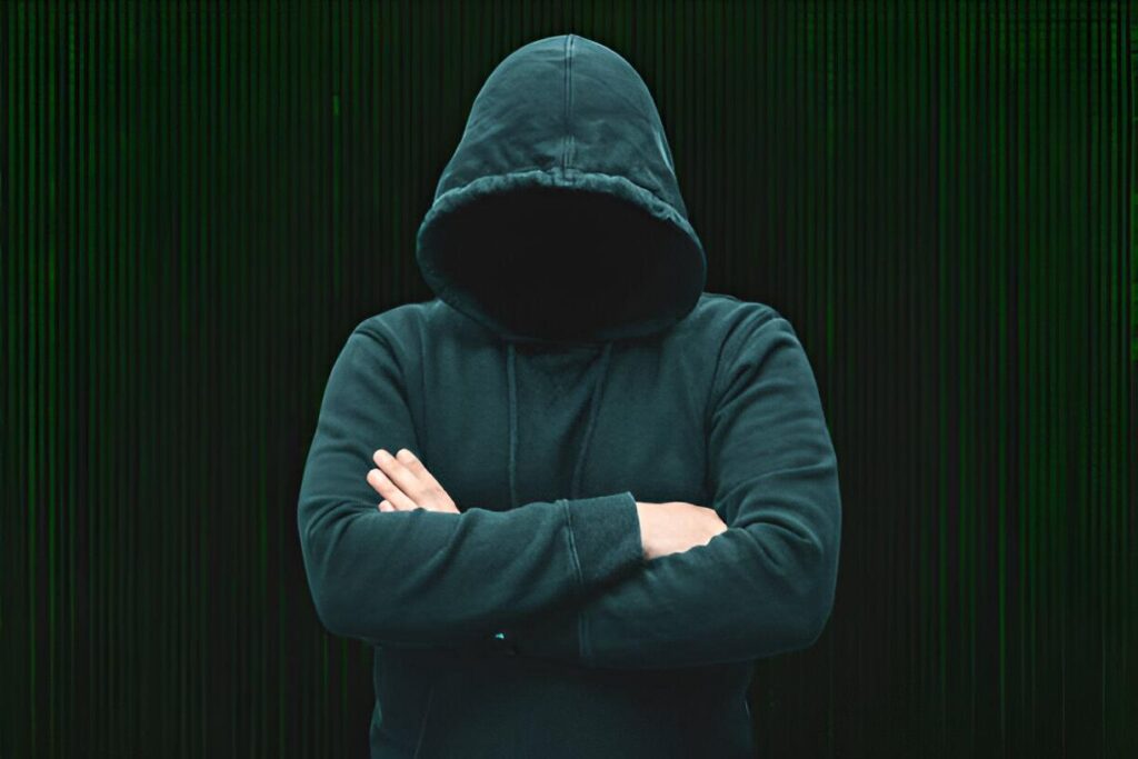 Alt text: A person wearing a dark hoodie with the hood up, obscuring their face, stands with arms crossed against a blurred dark background.