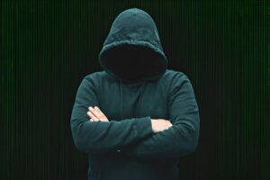 Alt text: A person wearing a dark hoodie with the hood up, obscuring their face, stands with arms crossed against a blurred dark background.