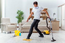 **Alt text:** A man energetically cleaning a living room by vacuuming and mopping simultaneously, with cleaning supplies nearby.