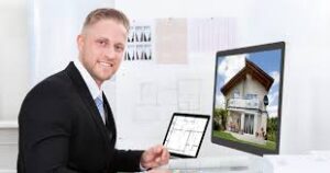Alt text: A man in formal attire sits at a desk with architectural plans on a tablet and a computer displaying a house design.