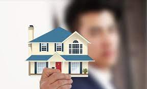 **Alt Text:** A person holds a miniature model of a house with a blue roof and a red door.