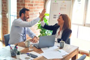 **Alt text:** A man and a woman in business attire exchange a high five at a desk with a laptop and papers in an office setting.