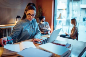 Alt text: A woman in glasses works on a laptop at a wooden table with books and papers, while two other people are seated in the background.