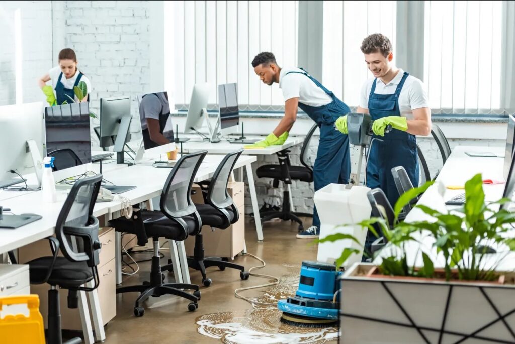 Alt text: A team of cleaners in uniform and green gloves working in a modern office, with one person polishing the floor, another handling a plant, and a third wiping desks near computer monitors.