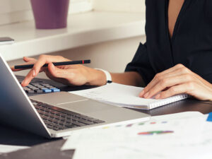 **Alt text:** A person using a calculator while taking notes beside a laptop on a desk.