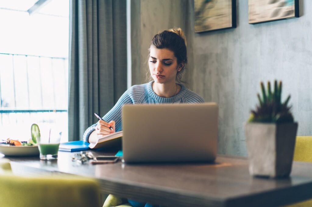 **Alt text:** A woman is sitting at a table, writing in a notebook with a laptop open in front of her, a plate of food, and a drink nearby.