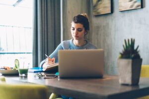 **Alt text:** A woman is sitting at a table, writing in a notebook with a laptop open in front of her, a plate of food, and a drink nearby.