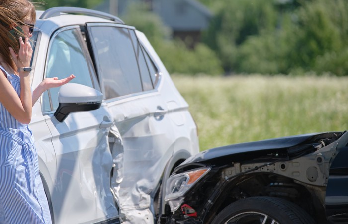 **Alt Text:** A woman stands beside a white SUV with a damaged side, while a black car is parked with its hood also damaged in a grassy area.