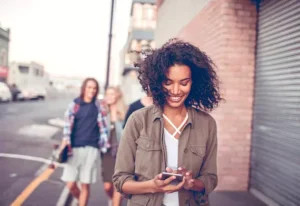 Alt text: A woman walks down a street looking at her phone, smiling, with two people walking behind her.