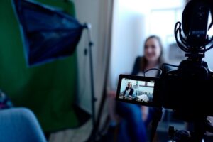 **Alt text:** A woman sits in front of a green screen being filmed by a camera during an interview setup.