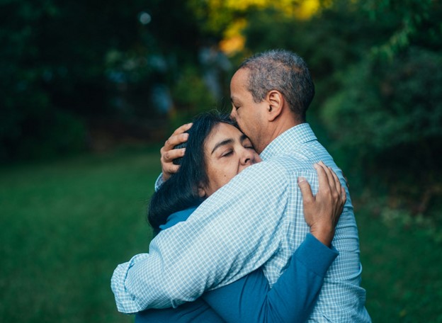 **Alt Text:** A couple embraces tenderly in a lush green outdoor setting, with the man gently kissing the woman's forehead.