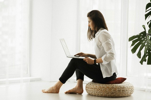 Alt text: A woman sitting cross-legged on a round cushion uses a laptop in a bright, minimalistic room with large windows and a plant.